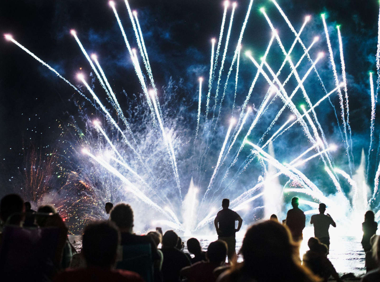 An audience watching a large, synchronized fireworks display over a lake at night.