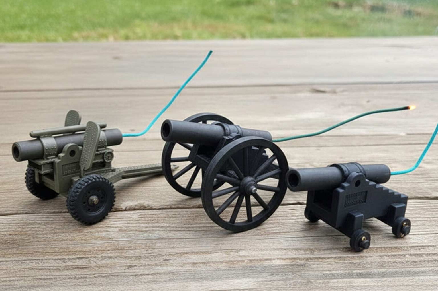 Three mini black powder cannons resting on a wooden surface with green Visco cannon fuse inserted into their vents, one with a lit burning end.