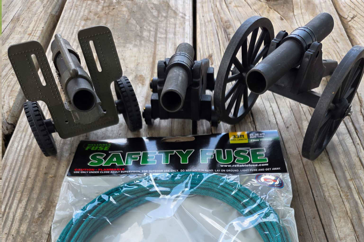 Three miniature black powder cannons lined up on a wooden deck above a packaged roll of 3.0 mm Reliable Fuse green Visco safety fuse.